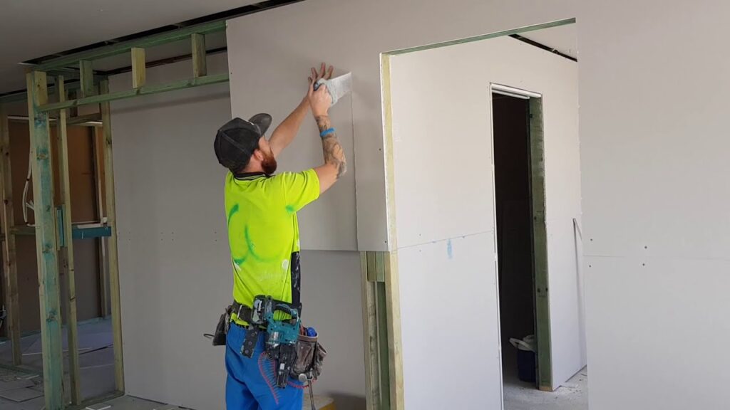A tradesperson in a bright yellow shirt uses a tool to apply joint compound to drywall in a room under construction.