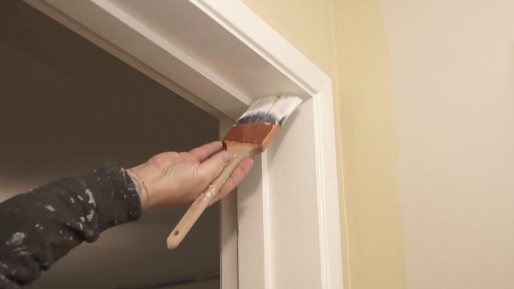 A hand using a paintbrush to apply white paint to a door frame molding.