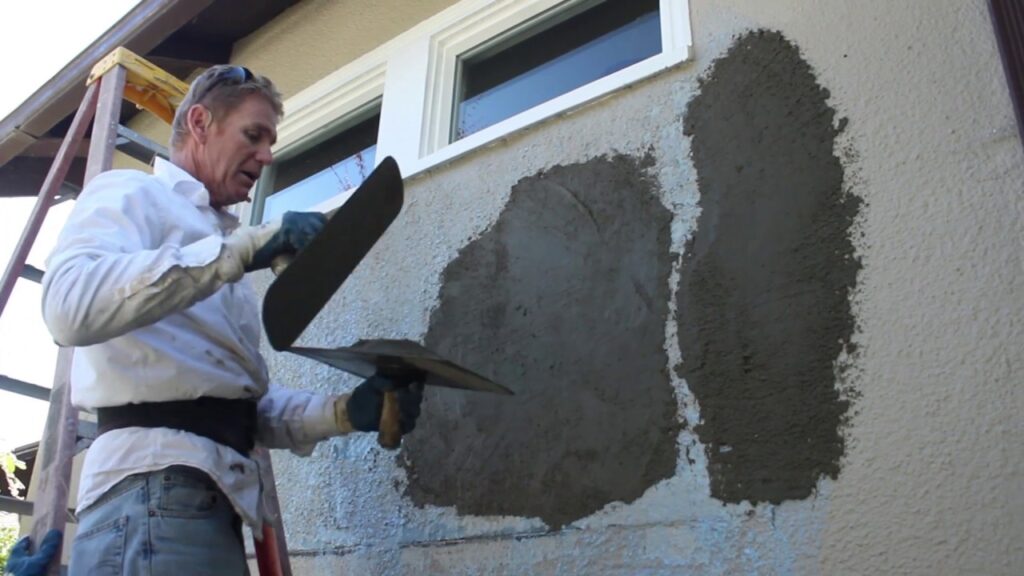 A man using a trowel and hawk to apply wet stucco patch to an exterior house wall.