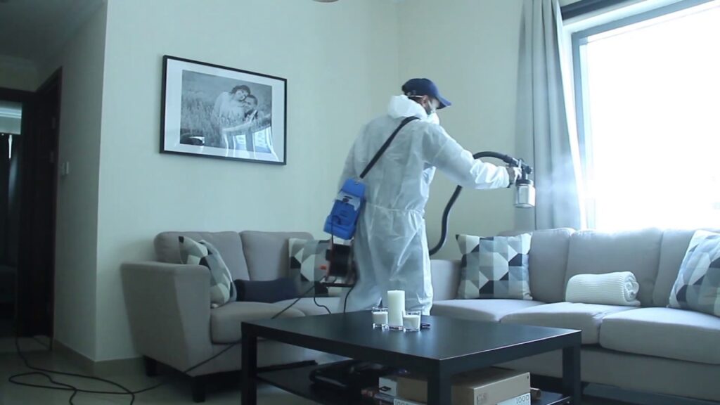 Person in a white hazmat suit spraying disinfectant mist inside a modern living room.