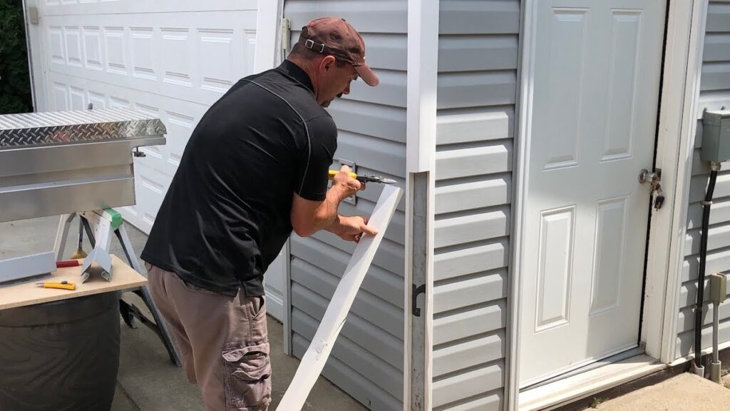 A man using tin snips to cut white corner trim on the exterior of a house with grey vinyl siding.
