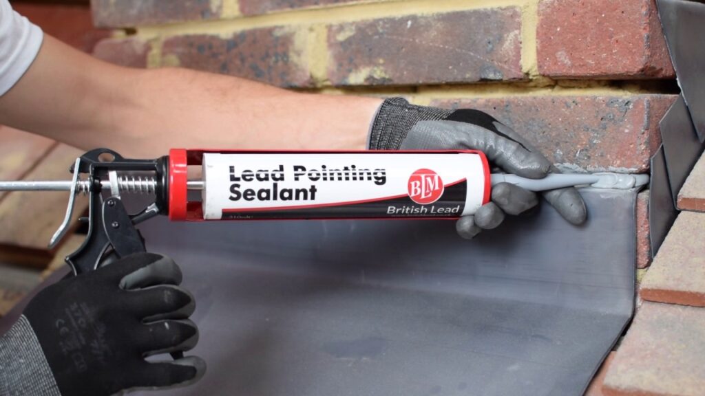 A person in gloves uses a sealant gun to apply Lead Pointing Sealant to a joint between lead flashing and a brick wall.