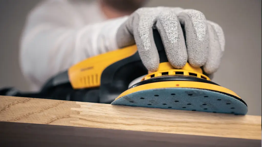 Person wearing gloves using an electric sander to smooth a piece of wood.