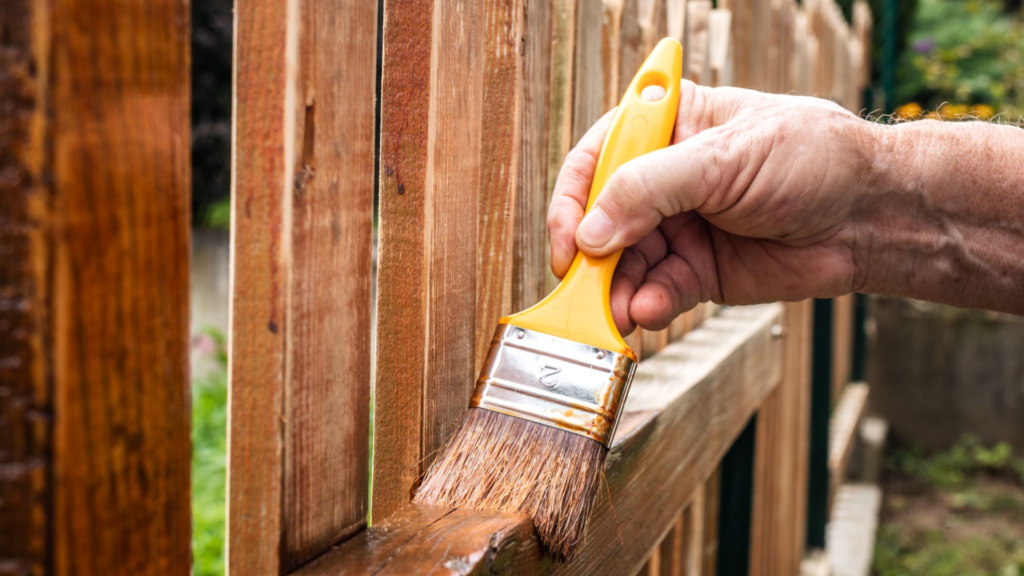A person's hand using a yellow brush to apply wood stain to a fence.