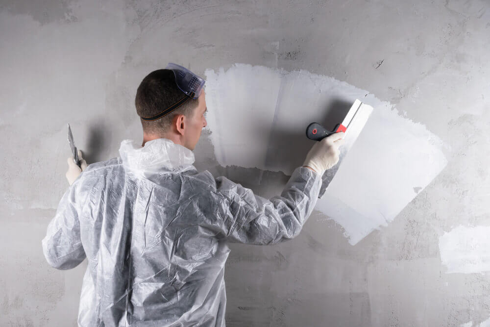 Construction worker in protective gear applying plaster to a grey wall with a trowel.