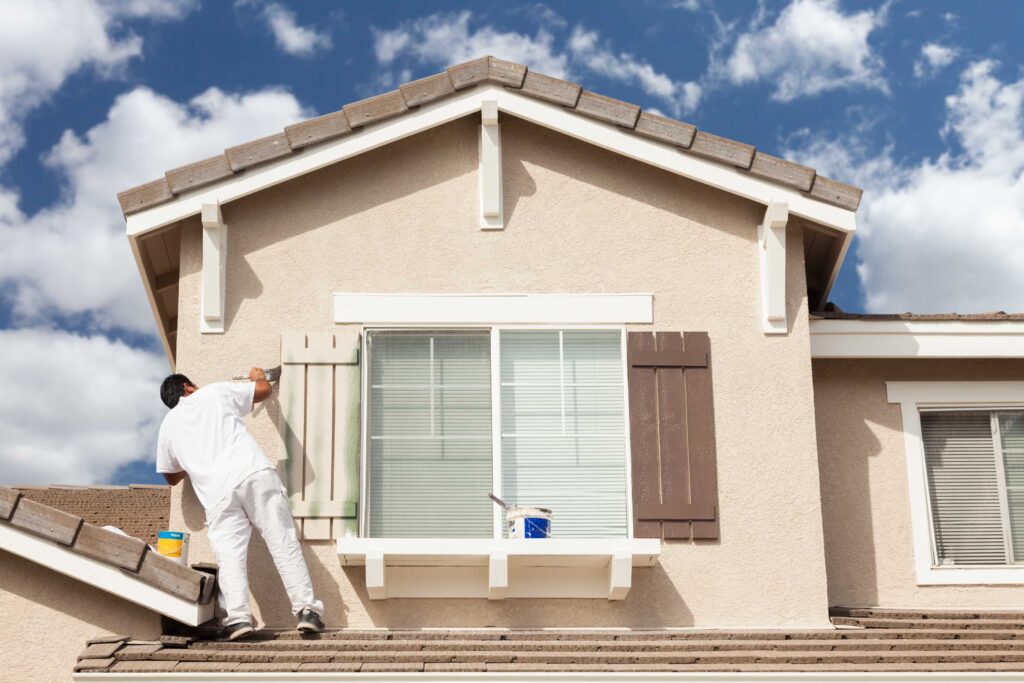 A house painter in white stands on a roof, painting a window shutter on a stucco home.