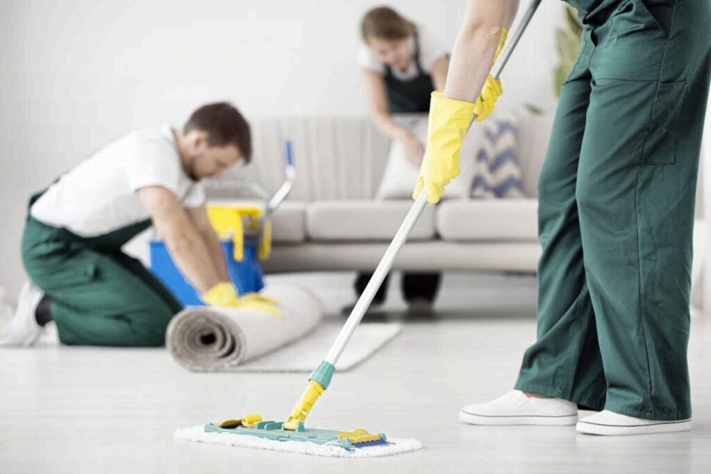 Three professional cleaners in green uniforms mopping the floor, rolling a rug, and cleaning a living room.