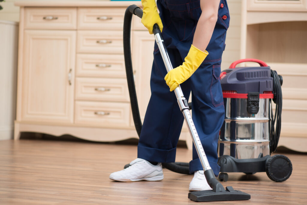 A person in blue overalls and yellow gloves uses a vacuum cleaner on a wooden floor.