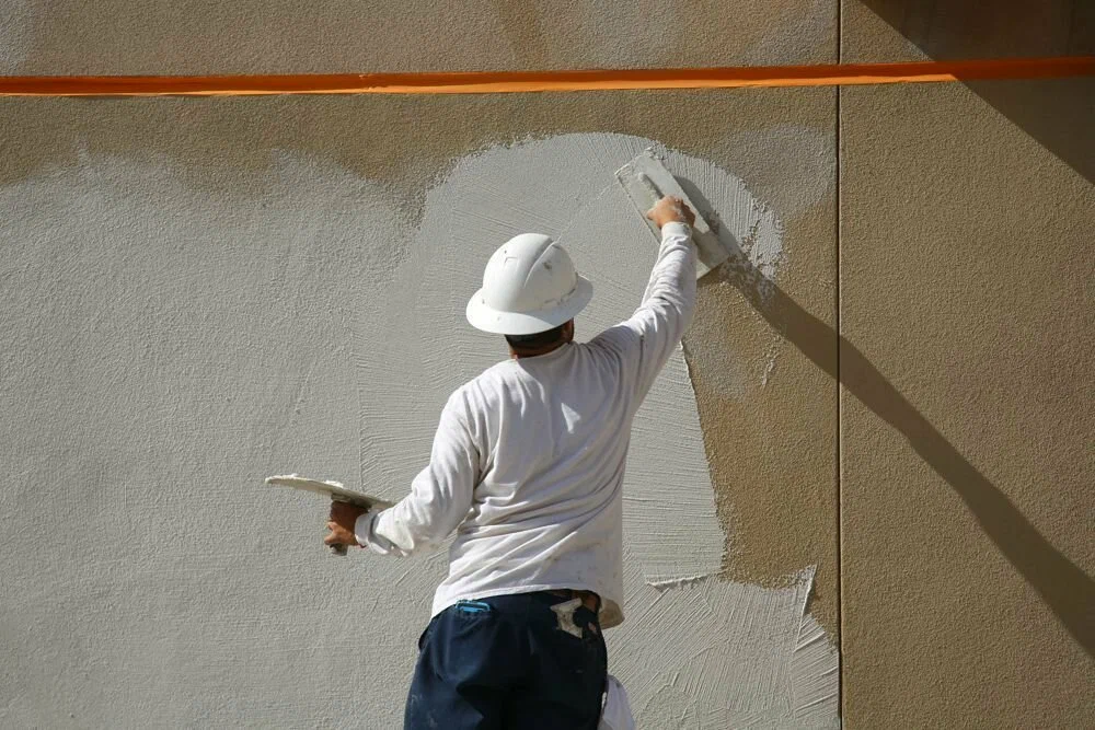Construction worker in a white hard hat applying stucco to a wall using a trowel.