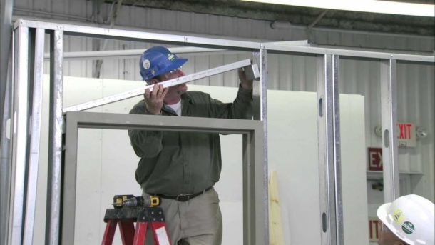 Construction worker in a blue hard hat installing a metal stud into a steel wall frame.