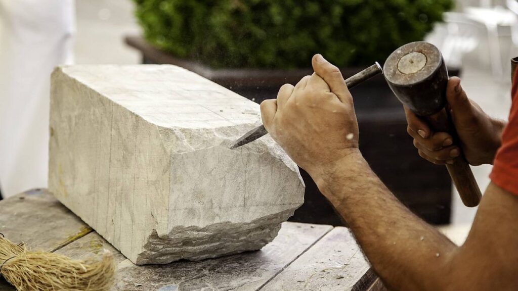 Close-up of a person's hands using a chisel and mallet to carve a large block of light-colored stone.