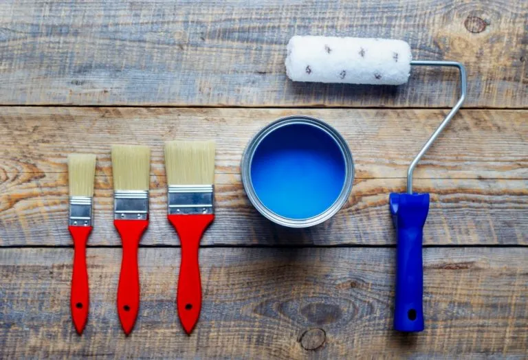 Three paintbrushes with red handles, a can of blue paint, and a paint roller on a wooden surface.