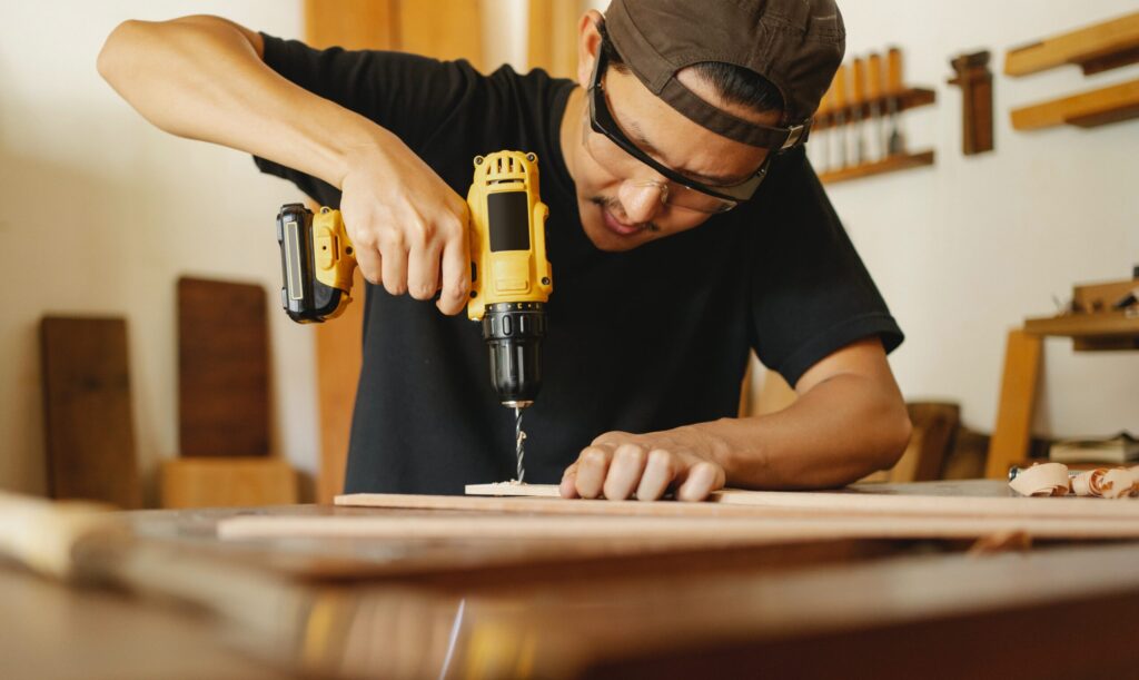 A man wearing safety glasses uses a yellow power drill to make a hole in a piece of wood on a workbench.