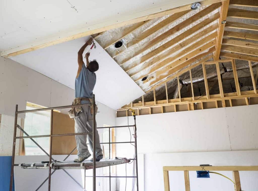 A construction worker standing on scaffolding installs drywall on a vaulted ceiling.