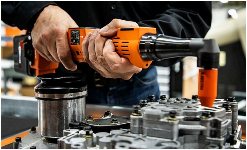 A worker uses an orange right-angle cordless power tool to assemble bolts on a metal mechanical component.