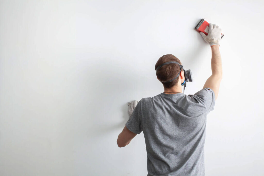 A man wearing a respirator and gloves sands a white wall with a sanding block.