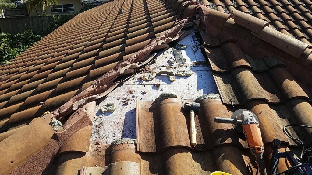 Broken terracotta roof tiles revealing underlayment and construction tools, including a hammer and angle grinder.