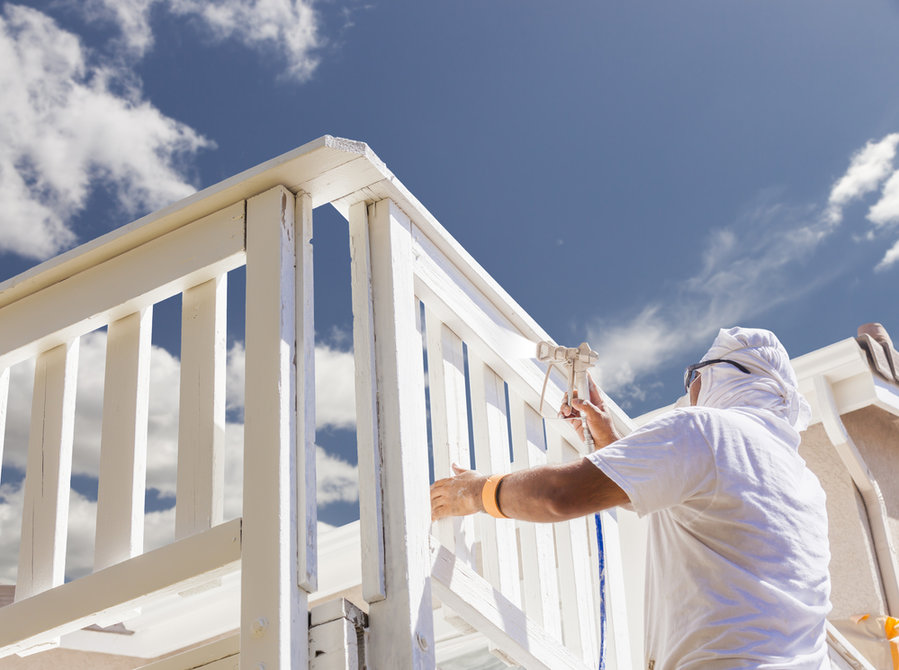 Painter in white protective gear using a spray gun to paint a white wooden railing against a blue sky.