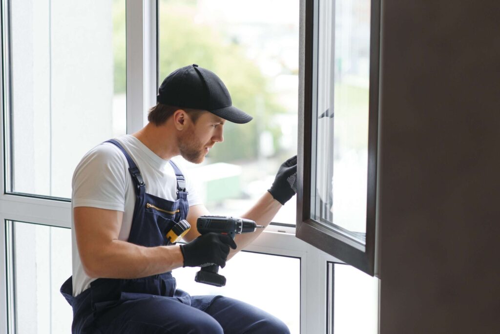 A technician wearing a cap and work overalls uses a power drill to install or repair a window.