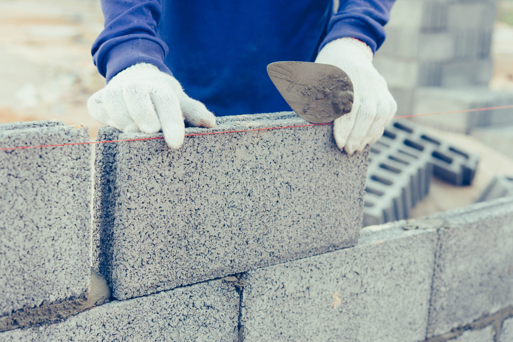 A construction worker in gloves places a concrete block on a wall while using a trowel and a leveling string line.
