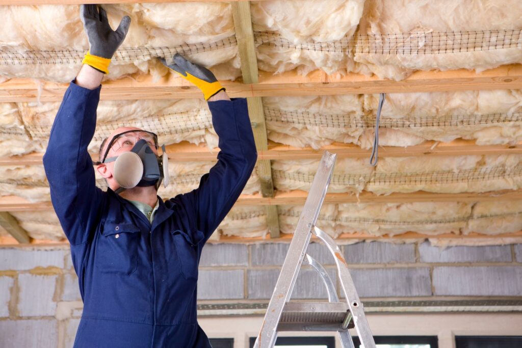Worker in blue overalls and a respirator installing fiberglass insulation between wooden ceiling joists.