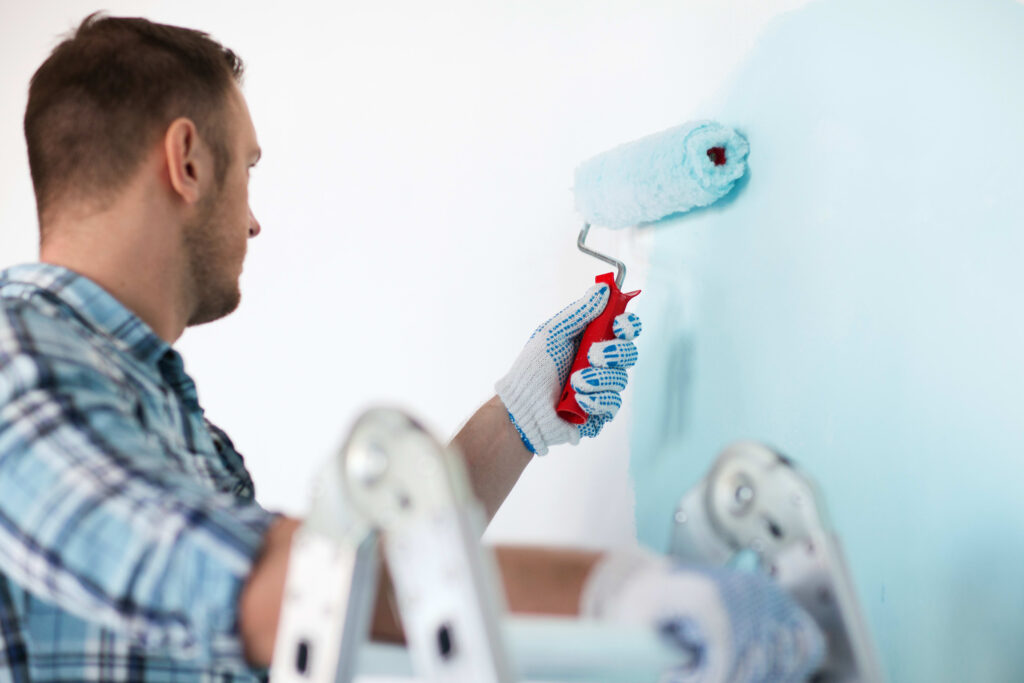 A man in a plaid shirt wearing protective gloves uses a paint roller to apply light blue paint to a white wall.