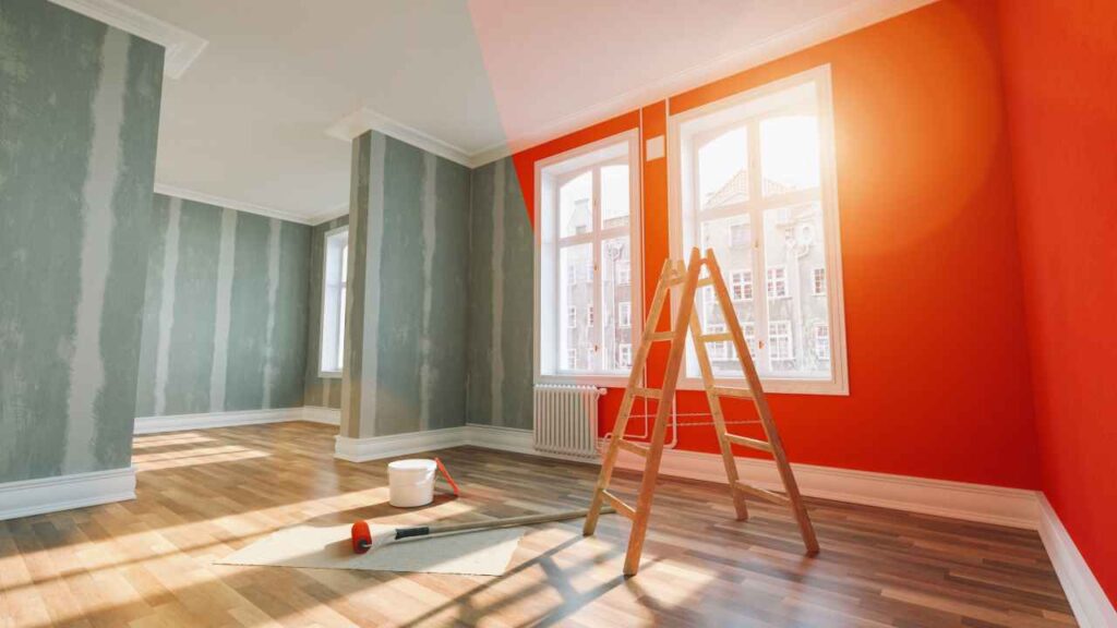 A room partially painted vibrant orange with a wooden ladder, paint bucket, and roller on the hardwood floor.