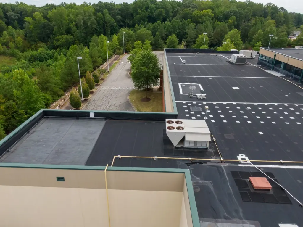 Aerial view of a commercial flat black roof with HVAC units, adjacent to a service road and dense green forest.