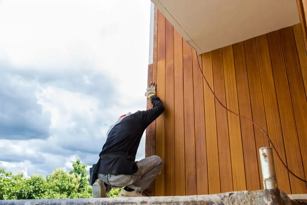 Construction worker installing vertical wood siding on a building exterior while crouching on scaffolding.