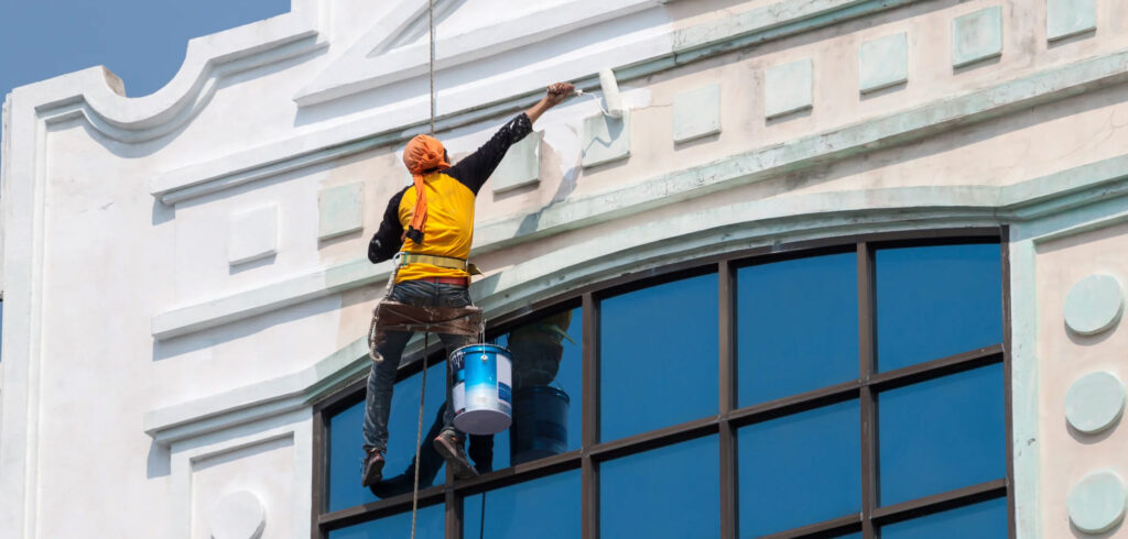 A worker suspended on a rope swing paints the white exterior of a building above large, blue-tinted windows.