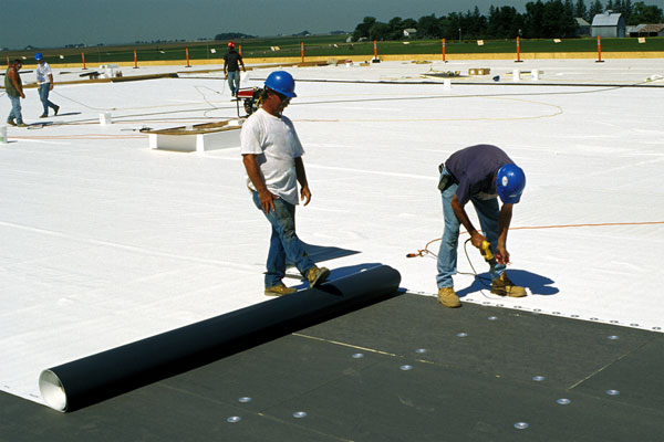 Workers wearing hard hats installing white roofing material on a large building site.