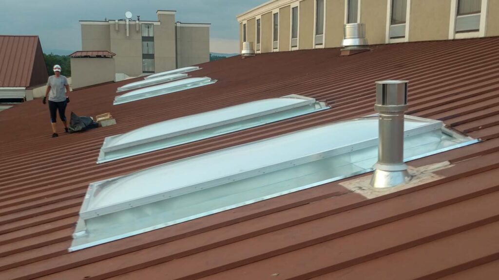 Worker on a brown corrugated metal roof with installed curved skylights and a silver vent pipe.
