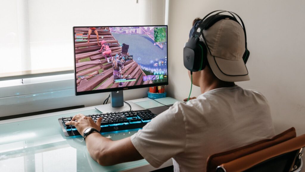 A person wearing a cap and headset playing a video game on a computer at a desk.