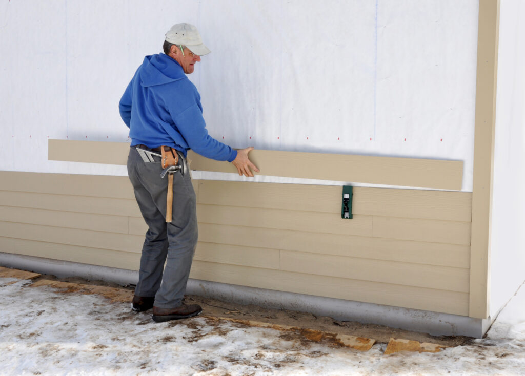 Man in a blue hoodie and tool belt installing tan siding on a house wall.