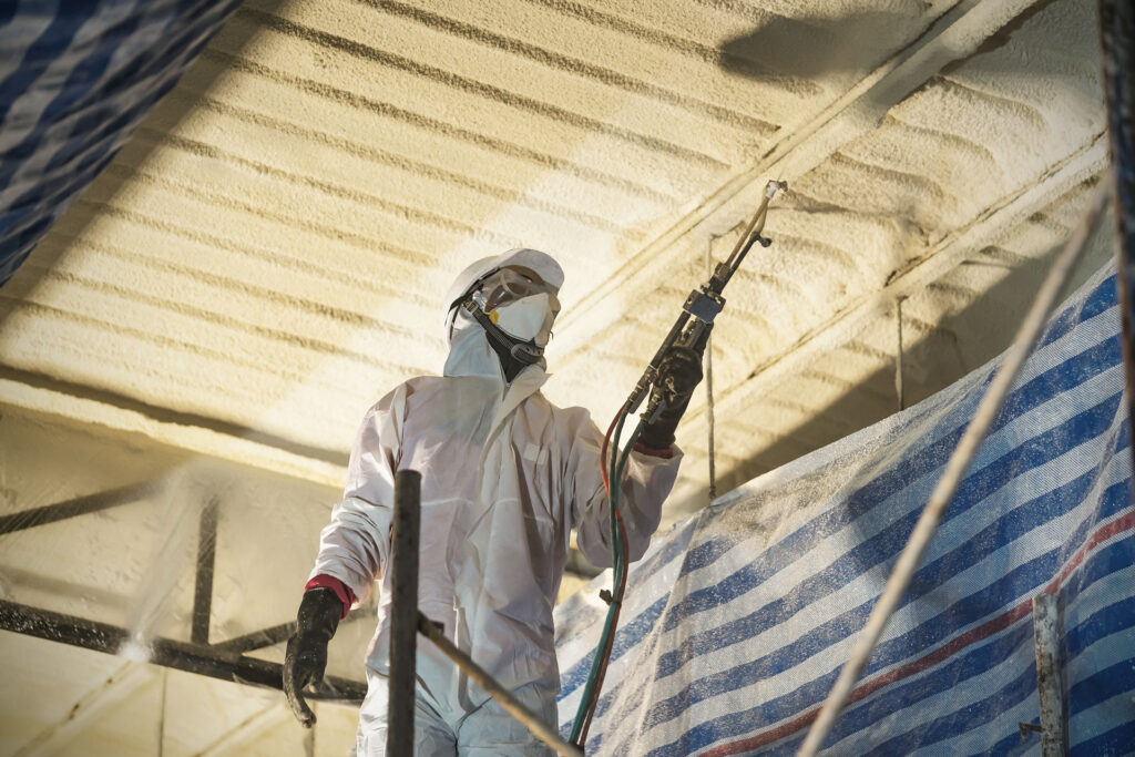Worker in a protective suit and mask spraying foam insulation onto a ceiling.
