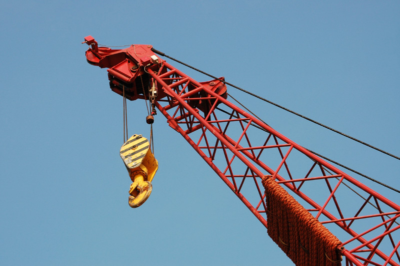 Red lattice crane arm with a yellow and black striped hook hanging against a clear blue sky.