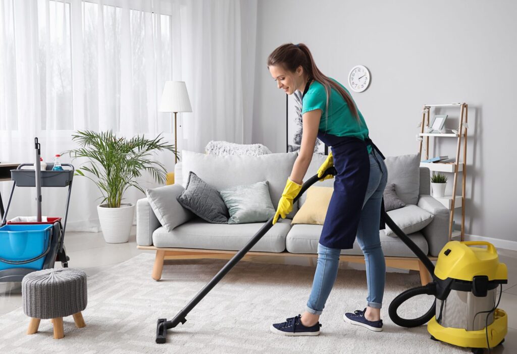 A professional cleaner in an apron and yellow gloves vacuums a rug in a bright, modern living room.