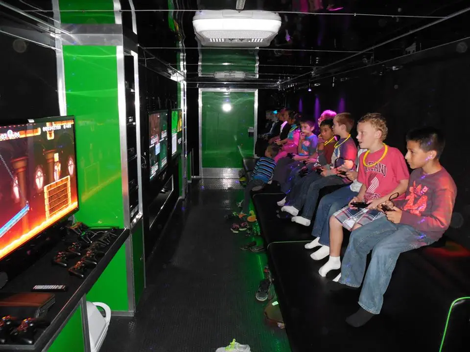 Children sit on a bench inside a mobile gaming trailer, playing video games on screens mounted to the wall.