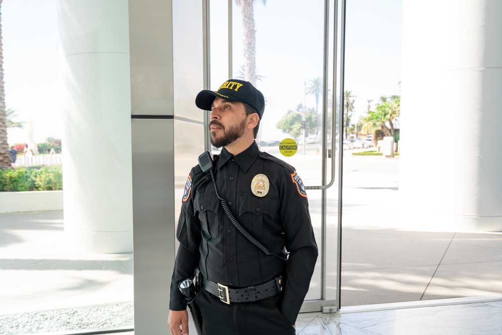 Security guard in a black uniform and cap standing near the glass entrance doors of a building.