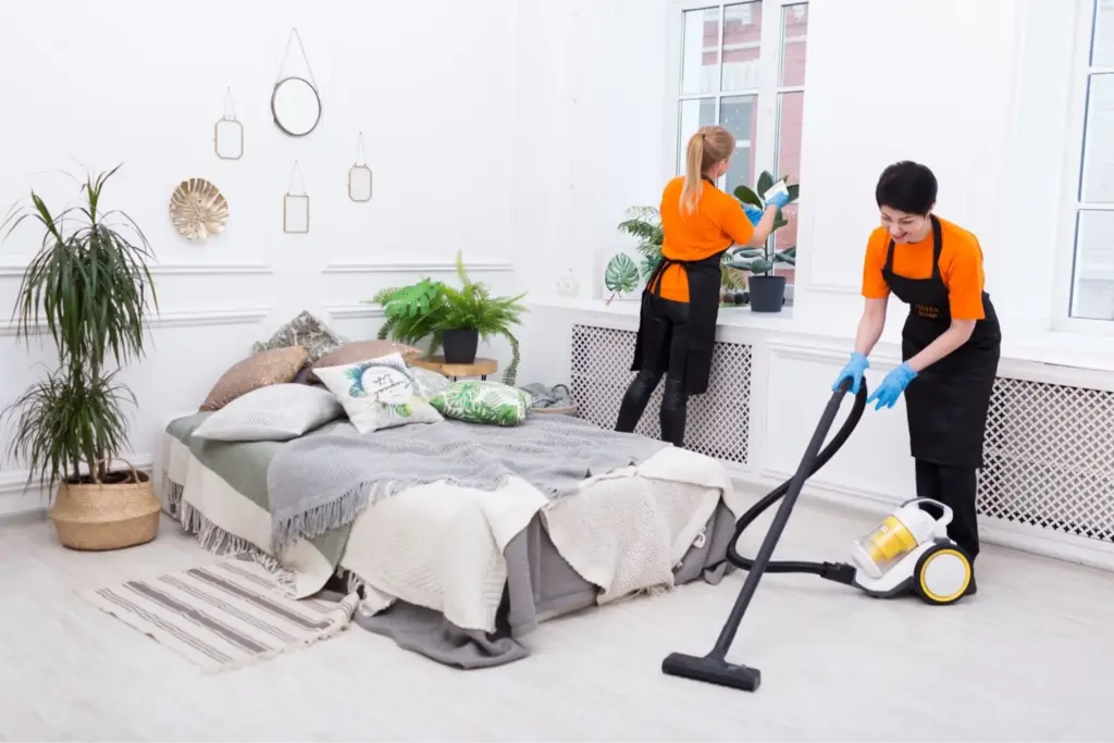 Two cleaners in orange shirts and black aprons vacuuming the floor and cleaning a window in a bright white bedroom.