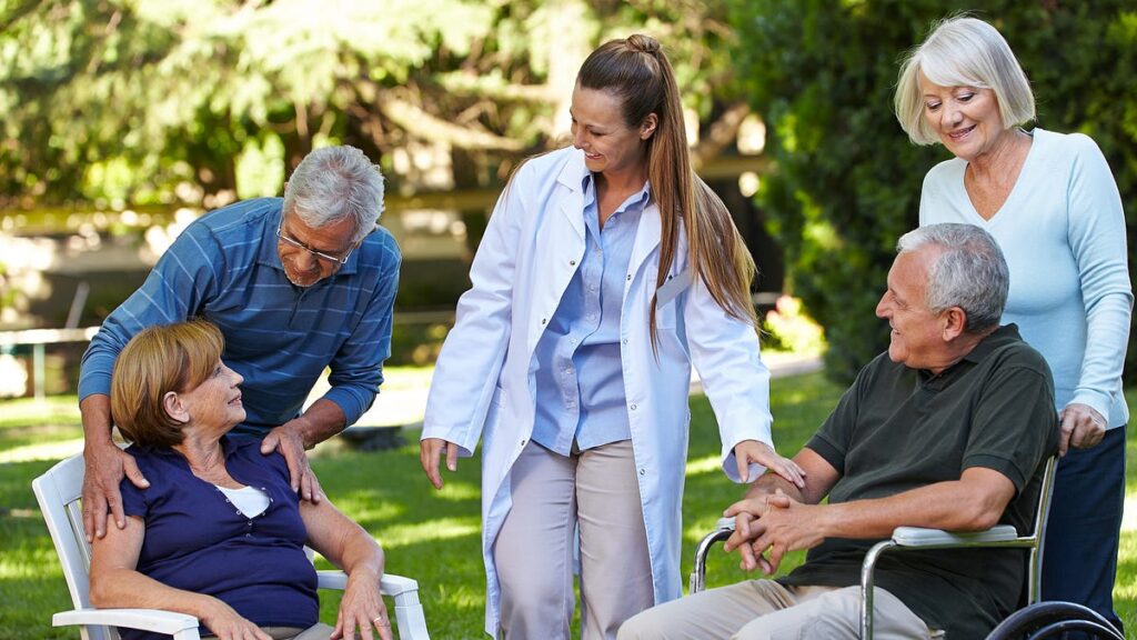 A female doctor in a white coat interacts with four senior citizens in a sunny outdoor garden.