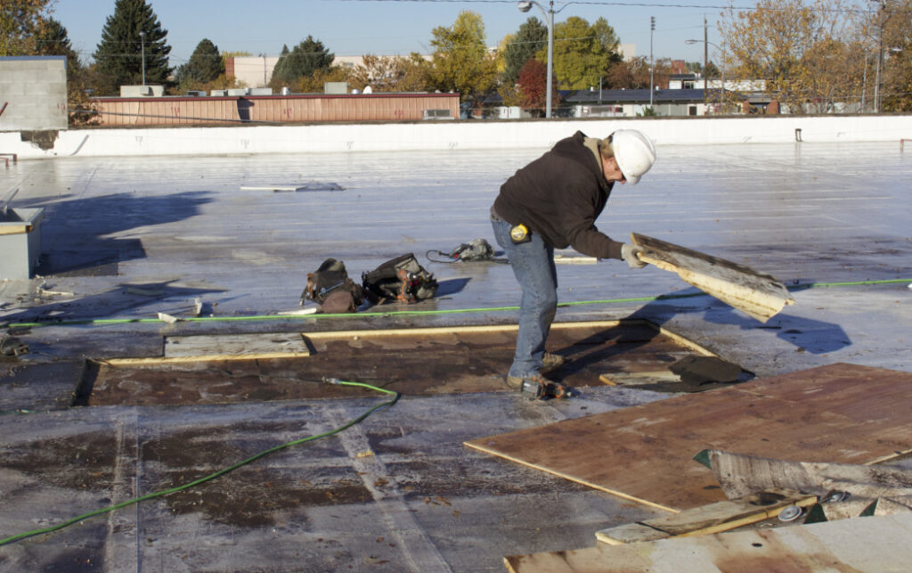A construction worker in a hard hat removes roofing material while repairing a flat commercial roof.