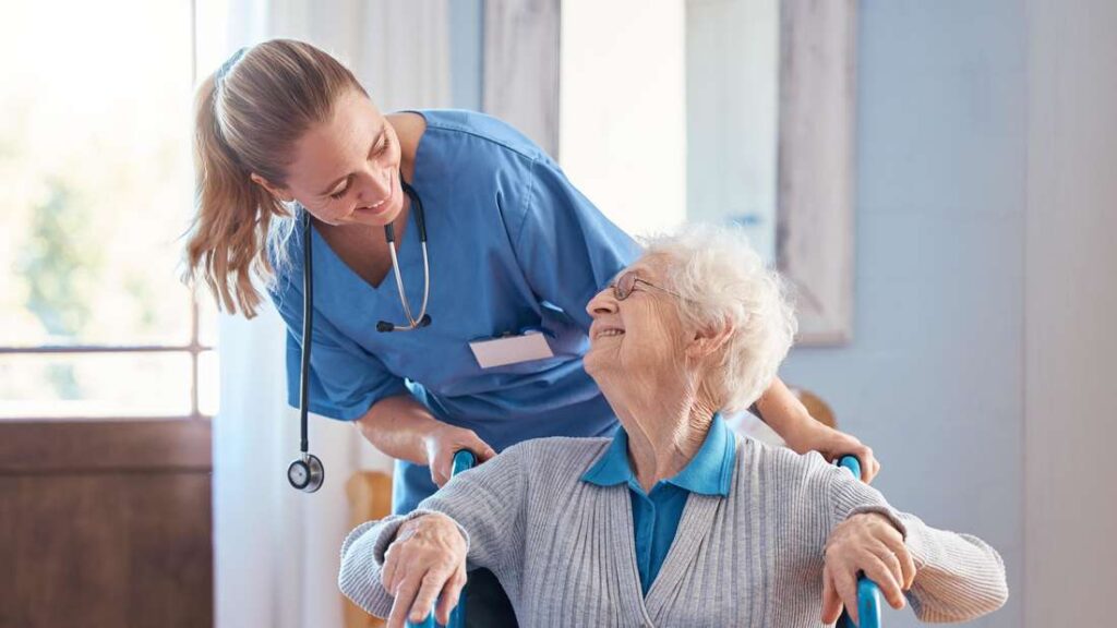 A smiling nurse in blue scrubs looks warmly at an elderly woman in a wheelchair, who smiles back.