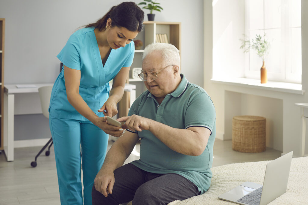 A nurse in blue scrubs helps an elderly man use a smartphone in a home care setting.