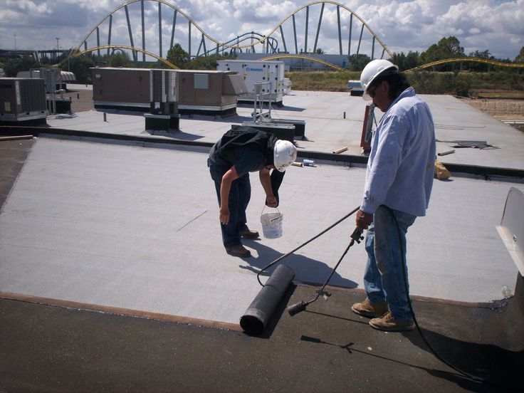 Two workers in hard hats install roofing material on a flat roof with a roller coaster in the background.