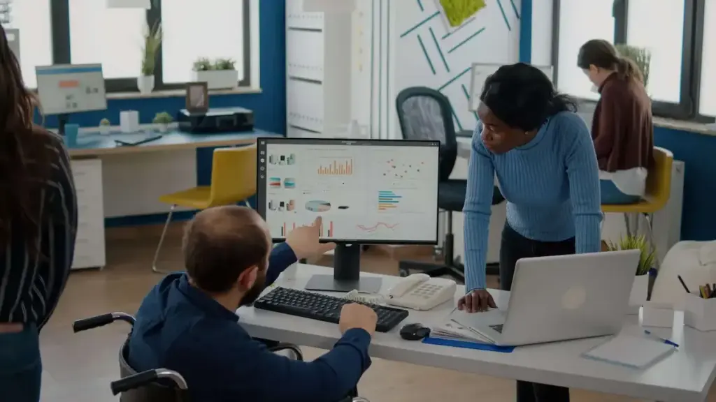 A man in a wheelchair points to data charts on a computer monitor while a woman in a blue sweater looks on.