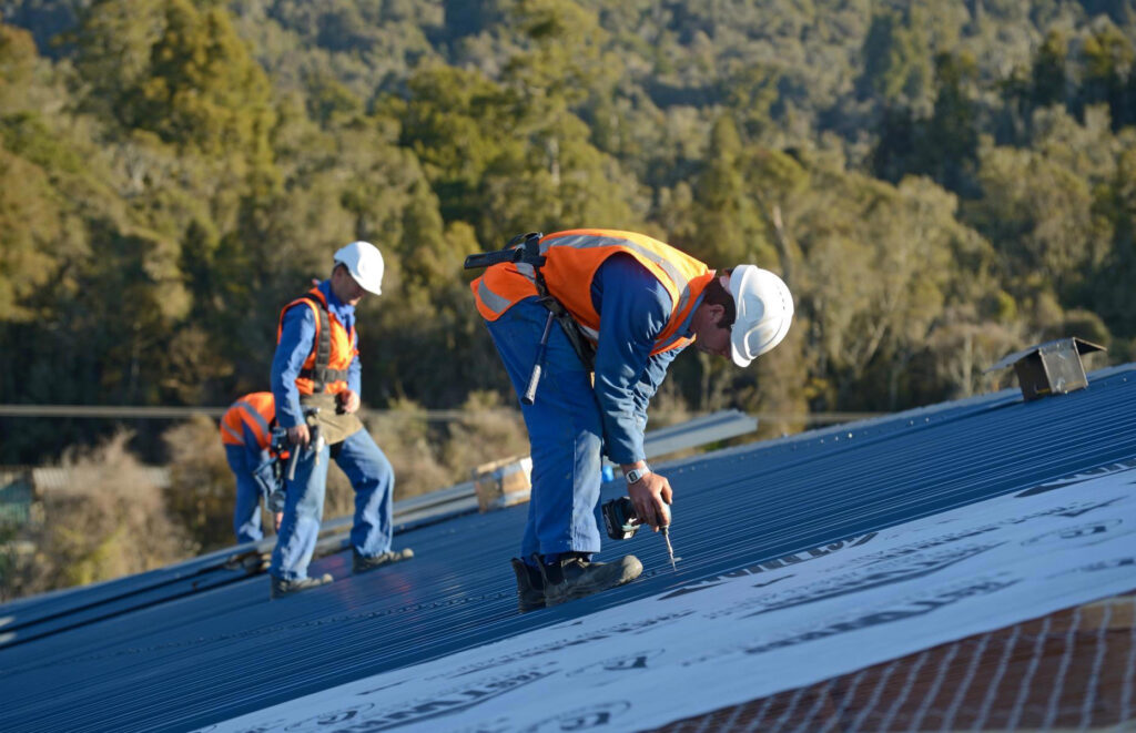 Roofers in safety gear install a blue corrugated metal roof, with one using a power drill.