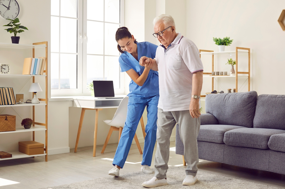A caregiver in blue scrubs helps an elderly man walk across a living room.