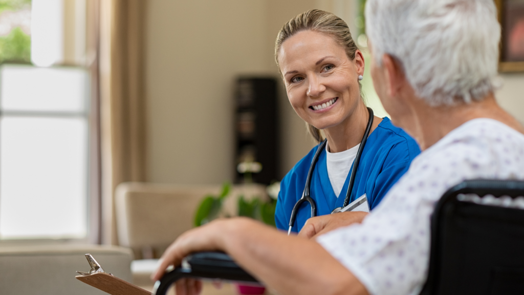 A smiling female nurse in blue scrubs with a stethoscope looks warmly at an elderly patient in a wheelchair.