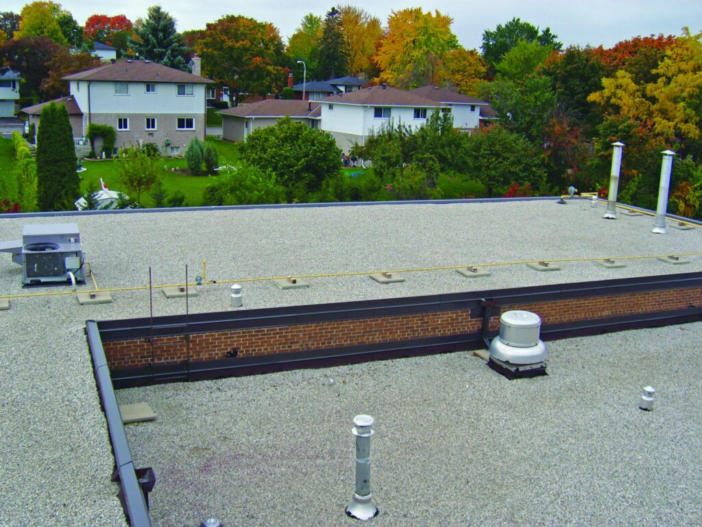 Flat gravel roof with HVAC equipment, vents, and a yellow gas line, overlooking a neighborhood with autumn trees.